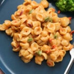 Tandoori masala macaroni and cheese on a blue plate with a fork and steamed broccoli to the side on a pink cloth.