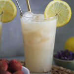 litchee juice lemonade in a glass topped with a lemon slice and lychees in a white bowl on the side.