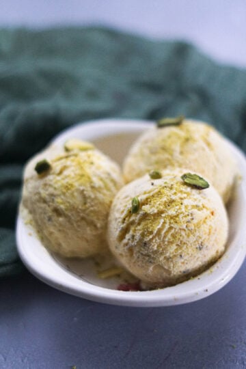Three kesar pista ice cream scoops in a small white bowl with a green cloth in the background.