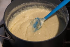 Tiramisu barfi mixture cooking on gas in pot with a blue spatula stirring it.