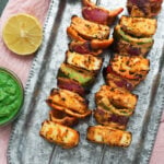 Grilled paneer tikka on skewers on a metal tray with chaat masala, green chutney and lemon to the side.