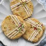 Three kesar pista cookies on a white fluted plate decorated with frosting and crushed pista and rose petals. on