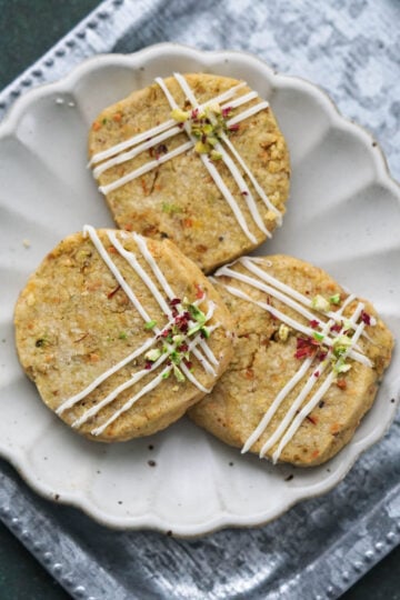Three kesar pista cookies on a white fluted plate decorated with frosting and crushed pista and rose petals. on