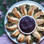 Samosa puff pastry wreath on a white platter with cranberry chutney in the center laying on a pink tablecloth.