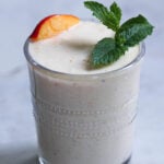 Peach lassi in a glass with a slice of peach and mint leaves garnish.