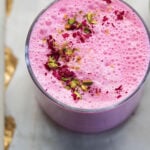 Rose lassi in a glass with pistachio and rose petal garnish.