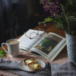 Chai with buttered toast and a book on a table with flowers to the side.