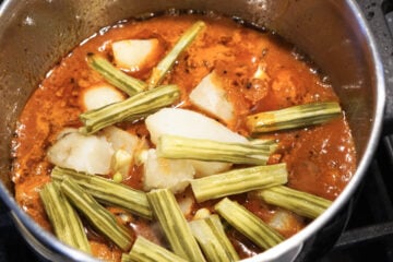 Potatoes and Indian drumstick vegetable added to tomato gravy in pot.