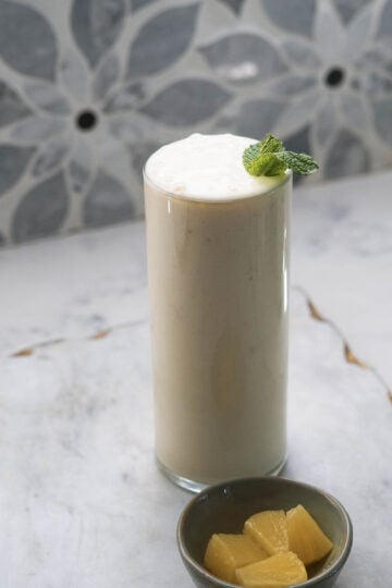 Tall glass of pineapple lassi with pineapple chunks in a small bowl to the side.