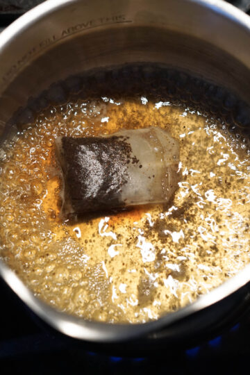 Black tea bag and chai spices simmering in water in a pot.