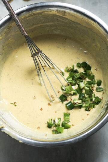 Besan batter and chopped green chilies in a steel bowl with a whisk.