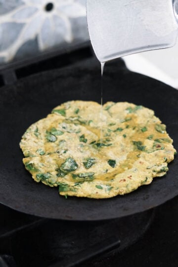 Drizzling oil on a thepla cooking on a tawa on stovetop.